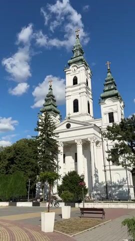 The video displays a static wide shot of a large, white, multi-towered church under a bright blue sky with scattered white clouds. The church features a central tall tower with a green, ornate spire and cross, flanked by two smaller side towers with similar green spires and crosses. Its facade has large white columns and arched windows. In the foreground, there is a paved plaza with decorative patterns, several green trees, white rectangular planters, and a brown park bench. Several individuals are seen moving across the plaza from left to right; one person in a yellow shirt is riding a bicycle, another in a red shirt is walking beside a bicycle, and a smaller figure is walking further back. The scene is well-lit by sunlight.