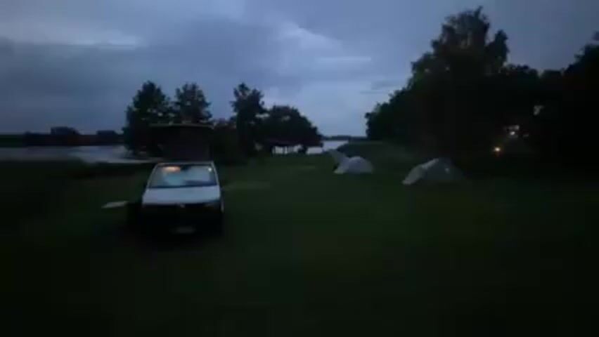 The scene is a dimly lit campsite at dusk. A white Volkswagen van is parked on the grassy field with its pop-up roof extended. Two tents are pitched in the background. Beyond the campsite, a body of water is visible under a cloudy sky. Trees and vegetation surround the campsite, adding to the natural setting.