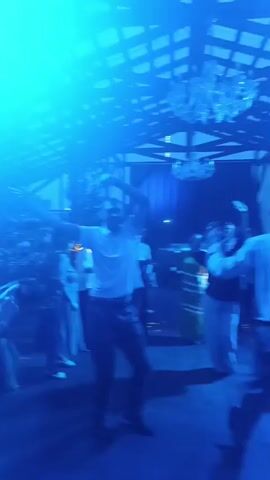 A man in a white shirt is dancing under blue lights. He has his arms raised above his head. Other people are blurred in the background, also appearing to dance. There is a chandelier hanging from the ceiling.