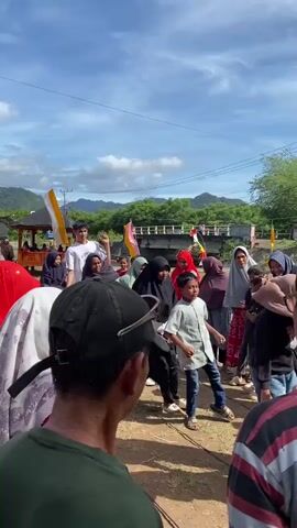 A group of people are gathered outdoors in a village. The weather is sunny with a blue sky and some clouds. In the foreground, the back of a man wearing a dark green t-shirt and a black baseball cap is visible. To his left, a woman in a red headscarf is partially visible. The main focus is on a group of people in the middle ground. A young boy in a light blue collared shirt and jeans is standing and looking towards the right. Several women are wearing hijabs in various colors, including black, green, and beige. Some are looking at the boy, while others are interacting with each other. In the background, there is a small wooden structure with a red roof, adorned with yellow and white flags. Further back, a bridge spans over a body of water, also decorated with flags. The landscape behind the bridge consists of rolling green hills. The ground appears to be dirt and gravel. The overall atmosphere seems festive or celebratory.