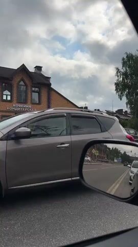 The video shows a view from inside a car, looking out the passenger side window and in the side mirror. The car is on a paved road, likely in a suburban or rural area. To the left, there is a brick building with a sign that reads "КОФЕЙНЯ ЦДИТЕРСКАЯ," suggesting it is a coffee shop and confectionary. Across the street from the building, there are parked cars, including a white van and darker colored SUVs. In the side mirror, the view is of the road behind the car, showing other vehicles in traffic. The sky is cloudy with patches of blue. Trees and houses are visible in the background. The overall impression is of a normal day of driving through a populated area.