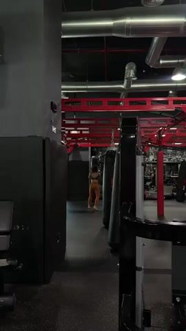 A person is seen in a gym from behind as they approach a punching bag. The person is wearing a sports bra and yoga pants. The gym has a black and red color scheme and a high ceiling with exposed pipes. Several punching bags are visible.