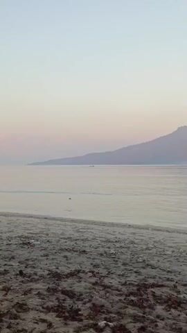 A drone shot pans across a beach and over calm water toward a distant, hazy mountain range. A small boat is visible on the water in the middle distance. The sky is a pale pink and orange, suggesting either sunrise or sunset.