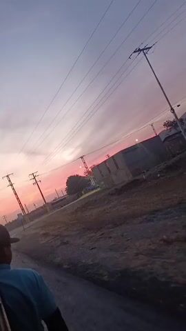 The video, captured from a moving vehicle, shows a rural landscape at sunset. The sky is a blend of pink, purple, and grey, with utility poles and lines silhouetted against the colorful backdrop. In the foreground, a person in a blue shirt is partially visible. As the vehicle progresses, a road sign indicating a bus stop comes into view. Across the road, there's a building and a blue bus parked. The ground is dusty and partially covered with vegetation. The overall scene suggests a quiet evening in a rural or industrial area.