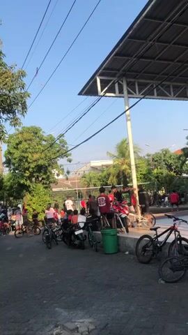 A group of people, mostly young men and boys, are gathered outdoors. Some are on motorcycles, while others are standing or sitting. Several bicycles are parked nearby. In the background, there is a volleyball net and what appears to be a covered outdoor area with a metal roof and supports. Trees and buildings are also visible. The sky is clear and blue. One person is holding a kite string, and a kite is visible in the sky. The overall atmosphere seems casual and social.