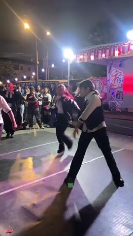 A group of young people are dancing on a raised platform at night. The platform is lit with green lights, and there are streetlights and stage lights in the background. A crowd of people are watching the dancers. The dancers are wearing black and white outfits. One dancer in the foreground is wearing a black jacket over a white shirt and black pants. Another dancer in the background is wearing a white shirt and black pants. They are performing a synchronized dance routine. There are colorful decorations and banners behind the stage. The overall atmosphere appears to be festive.