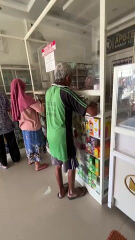 The video shows the interior of a pharmacy. Three people are visible. On the left, a person with short, light-colored hair is facing away from the camera, looking at shelves of medication. In the middle, a person wearing a pink hijab and a blue patterned skirt is also looking at the shelves. On the right, a man wearing a green t-shirt and shorts is standing at a counter, interacting with someone behind a clear plastic barrier. The shelves are stocked with various boxes and bottles of medicine. Signs are visible on the walls and the counter. The lighting appears to be artificial, with overhead lights illuminating the space.
