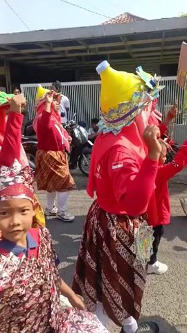 This is a video of a parade or procession on a sunny day. Several women, dressed in red tops, red headscarves, and patterned skirts, are walking in a line. They are all wearing distinctive conical yellow hats that resemble upside-down plastic bottles. Some of the women are carrying flags or small signs. The procession appears to be taking place on a street, with houses and buildings visible in the background. There are also motorcycles parked along the side of the road. Some onlookers are present, with at least one person holding a camera. The overall atmosphere seems festive and celebratory.