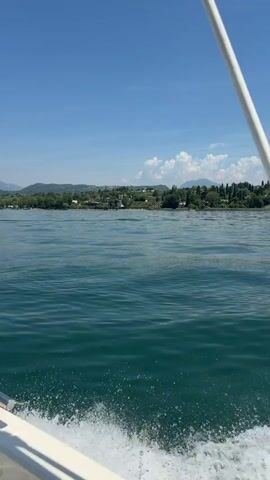 A vertical, first-person perspective video shows a boat moving through clear, blue-green water on a sunny day. The boat's white hull and a metal railing are visible in the foreground, with a spray of water trailing behind. In the distance, a green, hilly coastline is dotted with trees and houses, under a bright blue sky with scattered white clouds. The water has gentle ripples and appears calm.