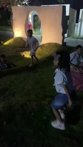 A group of children are playing on a grassy area at night. In the foreground, a young girl with dark hair and a white shirt and denim skirt is facing away from the camera and appears to be in mid-motion, possibly dancing or running. Behind her, another child in a light-colored dress is also in motion. In the background, there is a large, pale structure with an oval opening, illuminated from behind, and a child in a dark shirt is walking through it. The ground is covered in grass and some decorative mounds. Lighting appears to be from spotlights and ambient sources, creating a dark but illuminated outdoor scene.