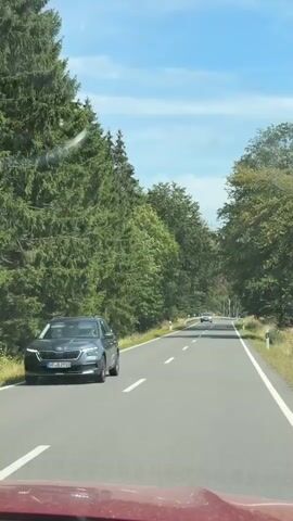 The video captures a first-person perspective of driving on a paved road through a forest. The road is two lanes wide with white dashed lines marking the lanes. There are tall green trees lining both sides of the road, with some taller trees and lighter foliage visible further into the forest. The sky above is blue with scattered white clouds. In the distance, a light-colored car is driving in the opposite direction, on the other side of the road. As the video progresses, the car comes closer and is identified as a gray Volkswagen ID.4 electric SUV. The video also shows a metal guardrail along the right side of the road and white posts with reflective markers at intervals. The sun is shining, casting shadows on the road. The overall impression is a scenic drive through a wooded area on a clear day.