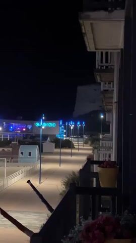 The video begins at night, focusing on a casino with blue neon lights. A walkway with multiple blue lampposts stretches from the casino into the distance. On the right side of the screen, a balcony railing is visible with a potted plant and some red flowers. The camera pans slightly to the left, revealing more of the walkway and a dimly lit building to the left of the casino. The video continues to pan left, showing the lights of the casino and the lampposts as they become more spread out, with a hill or cliff face visible in the background. The final shot shows a close-up of a lamppost with blue light emanating from it. There are no people visible in the video.