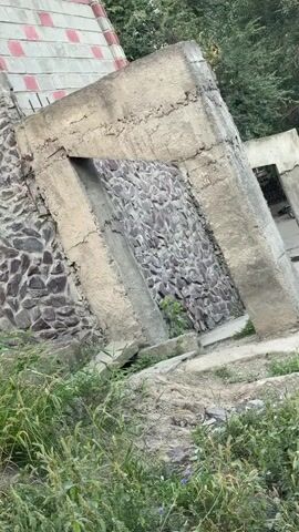 A video recording of an outdoor scene featuring an unfinished stone and concrete structure. The structure appears to be a wall with a large opening or doorway, made of rough stones and reinforced with concrete beams. To the left of the main structure, a building with a tiled roof and stone-clad lower section is visible. The surrounding environment is overgrown with green vegetation and grass. In the background, trees and a white truck are visible on a dirt road. The lighting suggests daytime.