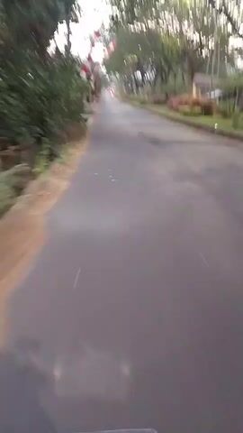 The video shows a view from a moving vehicle on an asphalt road. On the left side of the road, there is a concrete ditch, some greenery, and buildings with red and white striped flags. On the right side of the road, there are trees and some bushes. A dark-colored sedan is parked on the right side of the road. The sky is overcast. The video is slightly blurry, indicating movement.