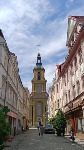 The video shows a street scene with a prominent church tower in the background. On either side of the street are buildings with shops on the ground floor and apartments above. The street is paved with cobblestones. There are a few people walking on the street and a car is parked in the middle of the road. The sky is blue with some clouds.
