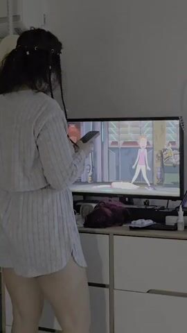 A woman with dark hair styled in braids with beads stands in front of a television screen, holding a black smartphone. She is wearing a light grey ribbed pajama set. The television displays an animated scene featuring a cartoon character in a pink outfit. The woman's posture suggests she is actively engaged with the content on the screen or the device in her hands. The background shows a dresser with various items on top, including what appears to be a dark red wig, a black cloth, and a bottle of lotion. The lighting in the room is dim, with the television screen being the primary light source.