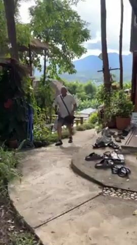 The video shows a man walking downhill on a paved walkway in a garden-like setting. He is wearing a white t-shirt, blue shorts with a design, and sneakers. He has a shoulder bag and carries a pair of black shoes. The walkway is bordered by lush greenery, including potted plants and trees. To the right, there are several pairs of sandals and shoes neatly arranged on a stone surface. In the background, there are small, thatched-roof structures resembling huts or gazebos, along with outdoor tables and chairs. Further back, a view of mountains and a town can be seen. The overall atmosphere is tranquil and natural, suggesting a resort or retreat setting.