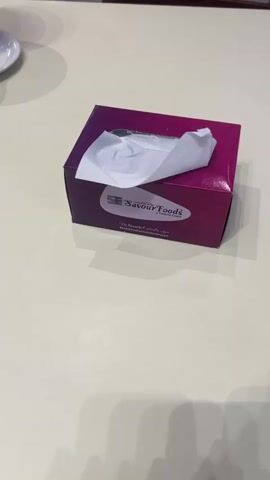 A tissue box with the Savour Foods logo is on a table. A piece of tissue is pulled out of the box. The camera pans over the tissue box.