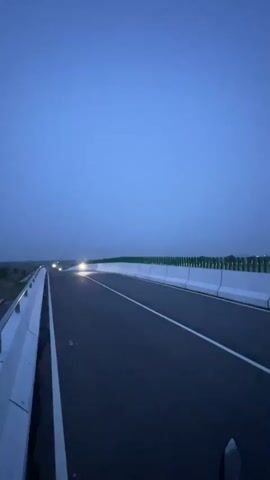 The video captures a motorcycle approaching and passing on an elevated section of a paved road at twilight. The sky is a uniform dark blue, indicating either dawn or dusk, and the motorcycle's headlights are illuminated. The road features clear white lane markings and is flanked by white concrete barriers on both sides. Beyond the right barrier, green fencing or vegetation is visible. The scene suggests an open highway or overpass, possibly in a rural or semi-rural area of Rupganj, Bangladesh. No other vehicles or people are visible, and no text is present.