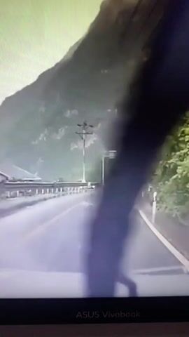 The video shows a first-person view of driving on a wet mountain road. The road has double yellow lines and white lane markings. There are guardrails on the left side of the road and a steep, tree-covered mountainside on the right. Signs are visible on the right side of the road, including an orange sign with yellow text and a blue sign with white text. There is a pedestrian crossing marked on the road. A few people are walking on the right side of the road, and a person can be seen riding a bicycle further up the road. The weather appears to be overcast and rainy, with visible raindrops on the windshield. The video is timestamped at the bottom, indicating it is from a recording.