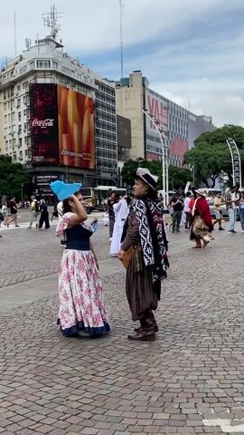 On a cobblestone plaza in a city, several people are dancing or preparing to dance, wearing traditional clothing. The plaza is surrounded by buildings with large advertisements, including a Coca-Cola ad showing French fries. A woman in a floral print dress waves a blue cloth above her head. A man in gaucho-style attire, including a hat, poncho, and brown leather chaps, faces her. Other dancers, some in white dresses and others in gaucho clothing, are nearby. People stroll around the plaza, seemingly watching the dancers. The sky is overcast.