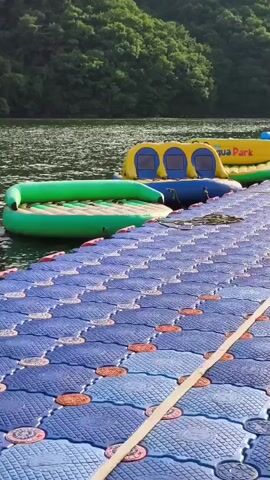 A floating dock made of blue interlocking modular plastic cubes with orange accents extends into a body of water. Adjacent to the dock are several inflatable water park structures, including a green circular inflatable with a raised edge, a yellow inflatable with blue sections and the words "Aqua Park" printed on it, and other colorful inflatables. In the background, a tree-covered hillside slopes down to the water's edge. The water appears calm with ripples. The sky is not visible.