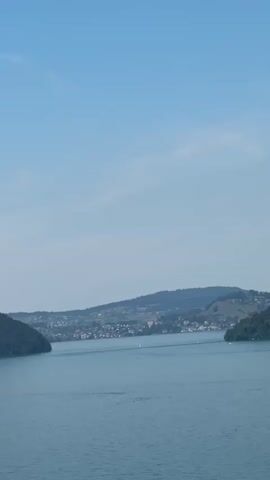 A wide shot of a large body of calm blue water under a clear blue sky with some wispy clouds. In the distance, rolling green hills dotted with houses and trees form a town along the shoreline. A narrow opening in the hills suggests a channel or bay. There are a few small sailboats visible on the water. The overall impression is a serene, natural landscape with human settlement.