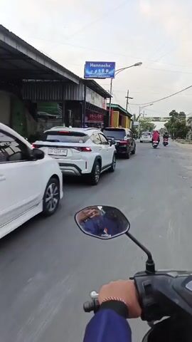 The video is shot from the perspective of someone riding a motorcycle on a paved road. In the foreground, the handlebars and a mirror of the motorcycle are visible. The mirror reflects the rider, who appears to be wearing a blue jacket and a watch on their left wrist. To the left of the road, there are several buildings with metal siding and glass doors, one of which has a sign with the text "Hannochs" and "DAMAR ELECTRONIC." Further down the road, there are other vehicles, including a white SUV, a black van, and several motorcycles. A person wearing a pink headscarf is riding one of the motorcycles. A large banner is strung across the road in the distance. The sky is overcast, suggesting it might be late afternoon or evening. The overall scene depicts a typical street view in a residential or commercial area.