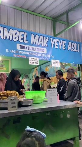 The video shows a food stall called "Sego Sambel Mak Yeye Asli" in Indonesia. A large blue banner above the counter states, "Tidak Buka Cabang," meaning "No branches," and lists the address as "Jl. Jagir Wonokromo Wetan No. 16." Several people are standing at the counter, appearing to be customers. A woman wearing a hijab is behind the counter, apparently preparing food. There are various bowls and trays containing food items on the counter. A sign indicates that fish ("Ikan Pari") is sold out ("Habis"). A menu is visible on the right side of the counter. There are signs indicating that customers can order takeout ("Antrian Bungkus") and that payment is made directly ("Pesan Langsung Bayar"). The stall has a green and white color scheme.