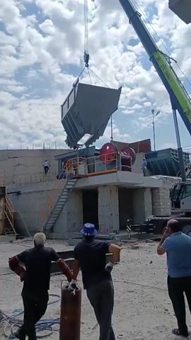 A crane is lifting a large gray hopper over a piece of industrial machinery that appears to be a crusher. The hopper is suspended by cables attached to the crane's boom. Below the hopper and machinery, three men are visible in the foreground. The man on the left is wearing a black shirt, dark pants, and protective gloves, and he is looking towards the hopper. The man in the middle is wearing a blue hat and a dark shirt, and he is also looking towards the hopper. The man on the right is wearing a blue shirt and dark pants, and he is holding his hand to his face. In the background, there are concrete structures, stairs leading up to the machinery, and another hopper visible in the distance. The sky is cloudy.