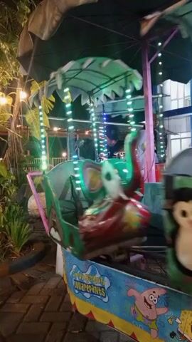 A young child wearing a light purple hijab and dress is seated in a colorful amusement park ride. The ride features various cartoon characters and is illuminated with string lights. The child is looking forward and appears to be enjoying the ride. The ride is a multi-unit contraption with each unit depicting different animals or characters. The background shows a dimly lit outdoor area with plants and some buildings. The video is shaky, suggesting it was recorded while moving.