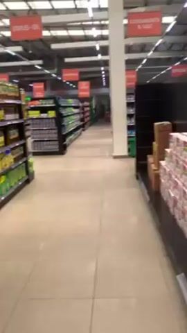 The video shows a walk through a grocery store aisle. The aisle has products like oil, condiments, and staples on shelves. The floor is a light tile. The lighting is bright overhead, and the ceiling is high. There are signs marking the sections of the aisle. No people are clearly visible in the immediate foreground. Boxes of Indomie noodles are visible on a pallet.