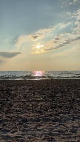 The video shows a beach scene with the sun setting over the ocean. The sand in the foreground is dark and textured, with small ripples and indentations. The ocean is calm with gentle waves, and the sun is a bright orb partially obscured by clouds, casting a shimmering reflection on the water. The sky is a mix of blue and pale yellow, with wispy clouds tinged with orange and pink. The camera is stationary, capturing the serene view. There are no people or man-made objects visible in the frame.
