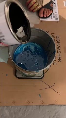 A close-up shot shows a person's foot, wearing a sandal and work glove, as they pour a dark liquid from a can into a bucket filled with blue liquid. The blue liquid is bubbly and shows swirling patterns as the dark liquid is added. The bucket rests on a piece of cardboard with some blue paint splatters and an 'X' drawn in marker. The background is slightly blurred but shows some packaging with text.