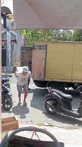 A person is carrying a box. The box is being loaded into the back of a yellow truck. The truck is parked on the side of the road. There are two motorcycles parked next to the truck. The person is wearing a white shirt and shorts. The person is wearing a blue head covering. There is another person in the back of the truck. The area appears to be an outdoor market or industrial area. The sun is shining and there are trees in the background.