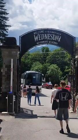 This video shows a wide shot of an entrance to what appears to be a filming location for the show Wednesday. Above the entrance is a large black arch with white lettering that reads "WELCOME TO THE FILMING PLACE OF WEDNESDAY NEVERMORE ACADEMY". Several people, appearing to be tourists, are walking through the archway. A tour bus is parked just inside the entrance. The background shows lush green trees and mountains under a bright blue sky with white clouds. The scene suggests a warm, sunny day. The focus of the video is the entrance arch and the activity around it.
