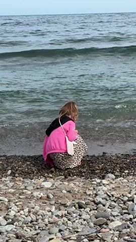 A young person with blonde hair, wearing a bright pink hooded jacket over a leopard-print skirt, crouches on a pebble beach near the water. They are looking down, seemingly observing something on the shore. A small white crocheted bag hangs over their shoulder. The ocean has moderate waves and the sky is overcast.