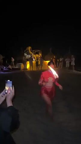 This media file depicts a nighttime performance on a beach, likely at Playa Del Sol-Villicana, Spain. The scene is dimly lit, with the primary light sources being a bonfire on the left, illuminated structures in the background, and spotlights on a stage-like area.

The central subject is a woman in a sparkly red costume, possibly performing a fire-dancing act. She is barefoot and appears to be twirling an object, possibly a fire staff, as evidenced by the flames visible on the right side of the frame. Her movements suggest a dynamic performance, and her expression is focused.

In the background, a group of people are gathered, observing the performance. Some are standing near a bar or food service area, indicated by tables and structures with lights. Others are positioned further back, watching the show. The presence of beach umbrellas with thatched roofs and sand suggests a tropical or Mediterranean beach setting.

The time of day is clearly night, with a dark sky. The weather appears calm, as the performance is taking place outdoors on the beach. The overall mood seems festive and engaging, with an audience present to witness the entertainment. The presence of a person holding up a smartphone to record the event indicates that this is a memorable occasion. There is no visible text within the media file itself.