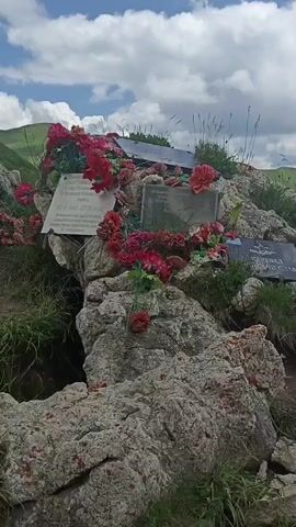 A video pans across a rocky, mountainous landscape. The scene features several memorial plaques and artificial flowers placed on the rocks, suggesting a site of remembrance. The environment is characterized by green grassy slopes, scattered snow patches, and a cloudy sky. The camera movement provides a panoramic view of the area, highlighting the natural beauty and the solemn nature of the memorial.
