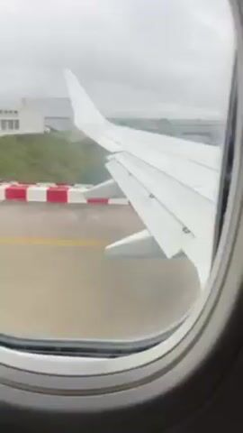 The view from inside an airplane window shows the white wing of the plane and a runway that it is taxiing on. The sky outside is overcast.  In the background are cars traveling on a highway.