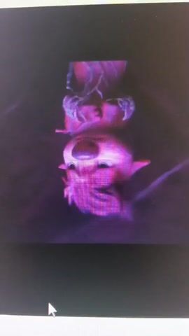 The video shows an animation of a character hanging upside down, suspended by thin lines, possibly strings. The character appears cartoonish with a large nose and stylized features, rendered in shades of pink and purple. Below the character, there's a glowing purple light source. The background is dark and blurred, with wispy, translucent patterns or effects. The image is displayed on a computer screen, and a cursor is visible in the lower left corner. The overall aesthetic is somewhat surreal and dreamlike.