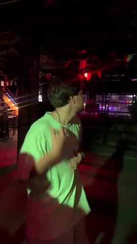 A young male with brown hair and wearing a white t-shirt and shorts dances in a dimly lit club. The lighting shifts rapidly between red, blue, and green. He moves his arms and body to the music, with a green wristband visible on his left wrist. The background shows a brick wall, bar area, and flags hanging from the ceiling.