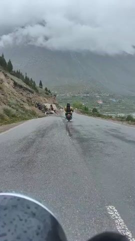 The video shows a paved road in a mountainous area, likely wet due to rain. A person on a scooter is riding away from the camera. On the left side of the road, a group of people in construction vests are working, with construction materials and debris visible. The background features a steep, tree-lined mountainside shrouded in clouds. The perspective suggests the video is filmed from another vehicle.