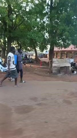 The video shows a dirt road in what appears to be a village or small town in a developing country. Several people are walking and riding motorcycles. There are simple buildings with red roofs along the side of the road. A few trees provide some shade. The sky is clear and the light suggests it is daytime. The overall scene appears to be an everyday moment in a rural area.
