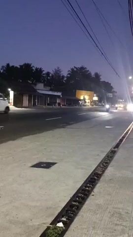 The video captures a street scene at dusk. A white van is traveling down the road, passing a row of small buildings on the left. A motorcycle with its headlight on is also visible on the road. Further down the road, a tow truck with its headlights on is visible, along with other vehicles. The sky is a deep blue, indicating twilight. Power lines are strung across the top of the frame. The ground is paved, with a drain grate running along the right side of the road. There are trees and vegetation visible in the background. The overall atmosphere is that of a quiet evening on a semi-urban road.