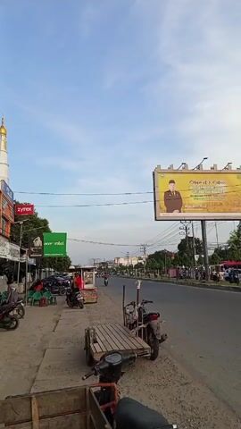 The video shows a view of a street in a city or town, likely in Indonesia, during daylight hours. The sky is blue with some light clouds. There is a two-lane road with traffic, including motorcycles and cars. On the left side of the road is a sidewalk with some parked motorcycles, a small food stall, and some computer stores. On the right side of the road are some buildings, including a medical faculty building. There is a large billboard on the right side of the road, which appears to be advertising a university. A large telecommunications tower is also visible in the distance. People can be seen riding motorcycles and walking along the sidewalk.