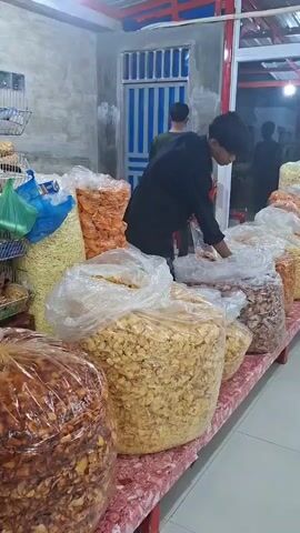 A young man in a black shirt, black pants, and wearing glasses, standing behind a long table filled with large transparent plastic bags of snacks, meticulously takes some of the snacks from a large bag and puts them into a smaller clear plastic bag. Another young man, wearing a green t-shirt and with short hair, stands behind him, observing. In the background, through a glass door, several people can be seen sitting around tables, also engaged in conversation or eating. The room is brightly lit, and the walls are a mix of light gray and white, with a striped blue-and-white barrier visible through a window. The snacks on the table come in various colors and shapes.
