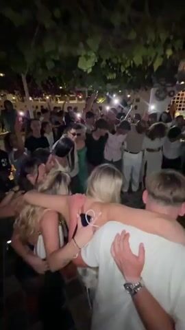 The video shows a crowded outdoor gathering at night. A group of people are huddled together in the foreground, with their arms around each other's shoulders, forming a circle. In the background, many more people are visible, standing and socializing. The environment appears to be a restaurant or bar with outdoor seating, as evidenced by the overhead greenery, lighting fixtures, and the presence of what looks like shelves or a bar in the far background. Some individuals are holding up their phones, possibly to record or illuminate the scene. The overall atmosphere seems festive and communal, with people engaged in a shared activity.