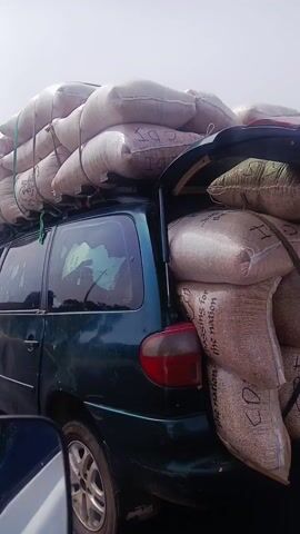 The video depicts a green minivan overloaded with large white sacks, secured with ropes on its roof. The van appears to be driving on a road with trees and buildings in the background. The video seems to be taken from inside another vehicle, showing a person with dark skin wearing a blue shirt and a white baseball cap with the letters "J-K" on it, looking at the camera briefly. The video then pans back to the outside, continuing to show the overloaded van driving down the road.