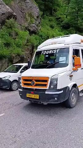 The video shows a white tour bus with "TRAVELLER" written on its side. The bus is driving on a road next to a rocky, green mountainside. A person in a yellow shirt is visible in the foreground. The bus is seen from the front, side, and rear. There are other vehicles and road barriers visible in the distance. The overall environment appears to be a scenic, mountainous region.